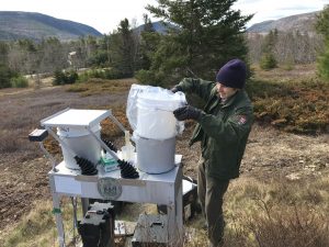 A technician replaces a plastic bucket on a metal table with trees and hills in background.