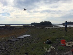 Stefan flies a drone over rockweed-covered shore. Calibration panels, square frames, and other gear in forefround.