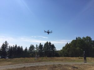 A pilot flies a drone from a gravel driveway with trees in background