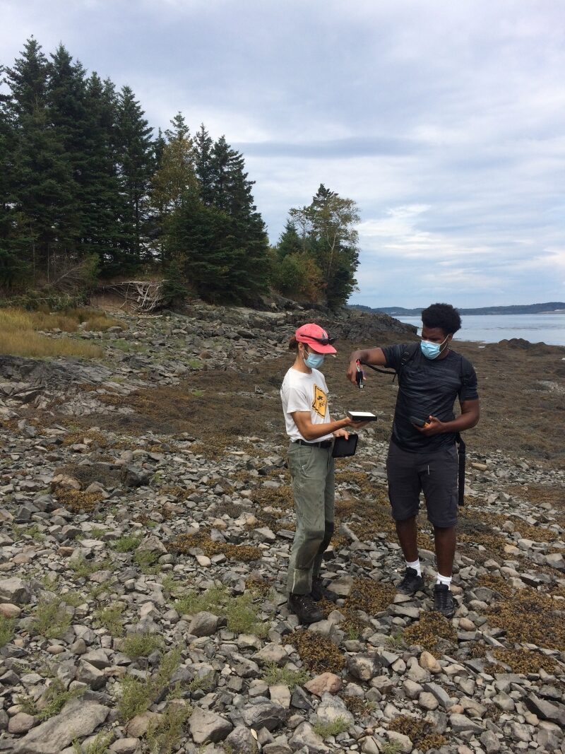 Two people stand on shore. One holds a calibration color and the other takes a reading.