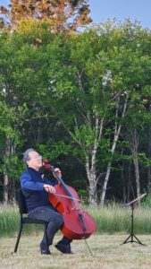 Yo-Yo Ma seated in a chair plays cello with grass, birch and spruce trees in background.