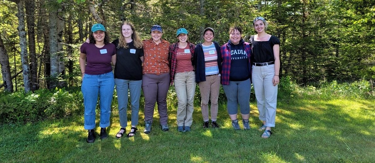 Seven of the nine Schoodic Institute techs and interns stand in a line with trees behind them