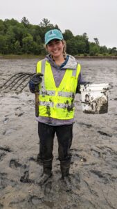 A muddy Elizabeth Halasz stands in the mudflat with clam rake and bucket.