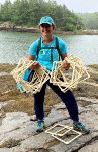 Jess Moskowitz stands on rocks in the intertidal zone holding sampling quadrats with water and trees in background.