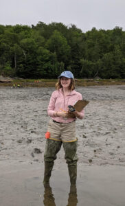 Anna Lee holds a clipboard and stands in shallow water with mud and trees in background.