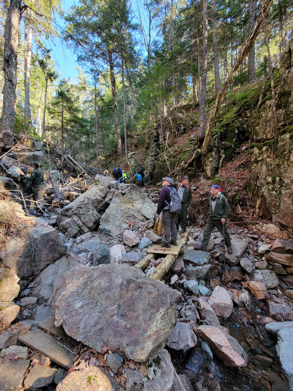 Three people stand in the ravine of Maple Spring. A large boulder sits on a broken wooden footbridge. Other large rocks and boulders fill the stream bed. Eroded banks are visible. More people examine damage in the background.