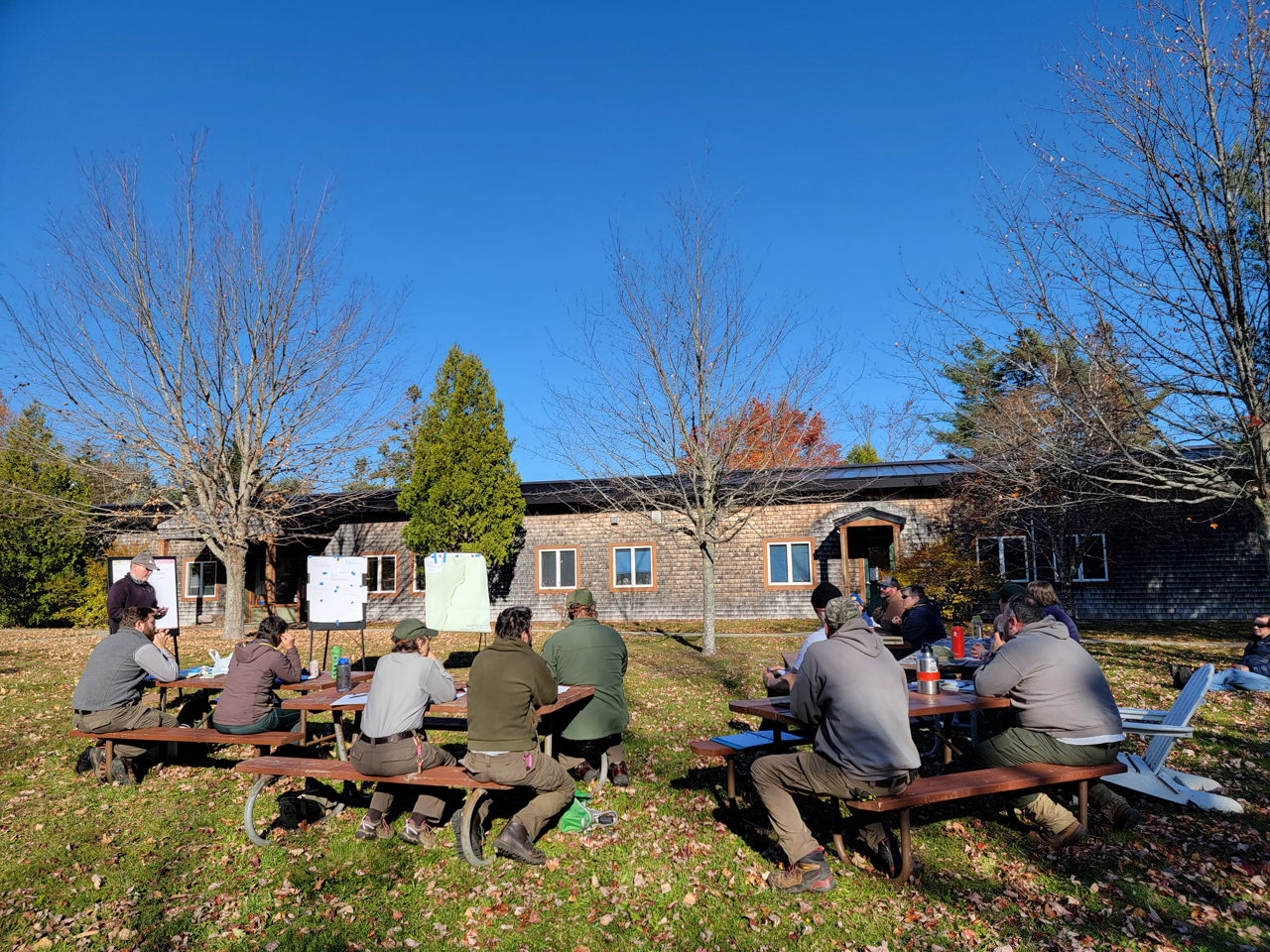 On a green lawn strewn with fallen leaves, a group of people, some in National Park Service uniforms, sitting at picnic tables in a half-circle, facing three easels where a person stands taking notes. The cedar-shingled park headquarters building and trees are in the background.
