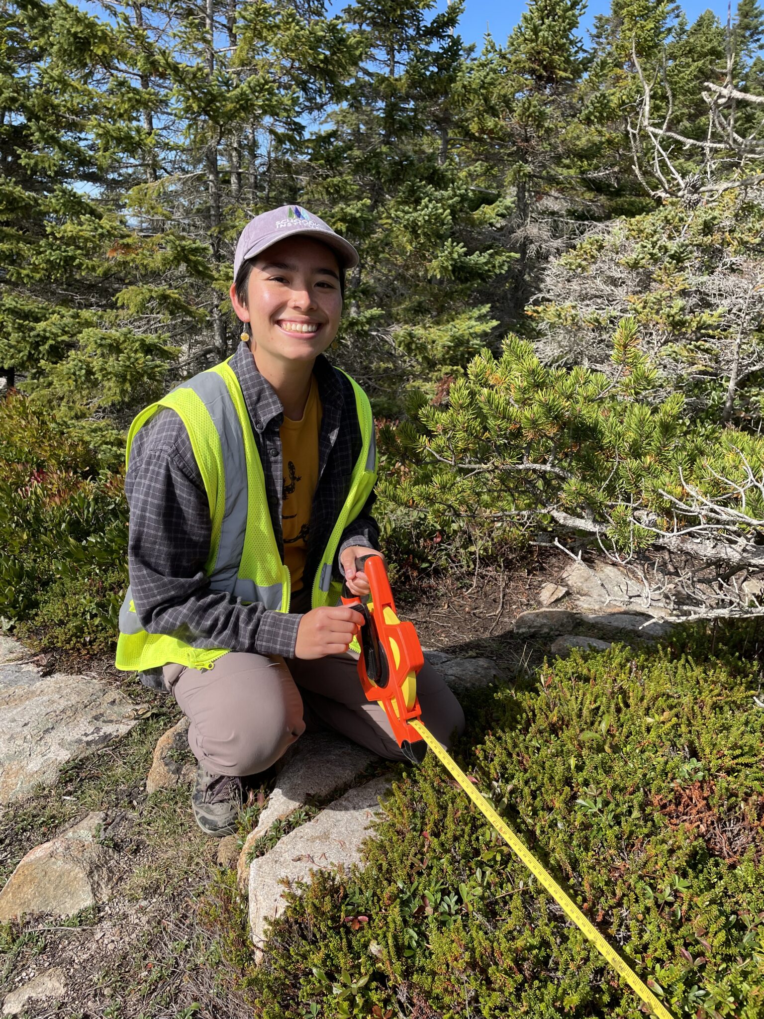 A smiling person kneels next to crowberry with trees in background.