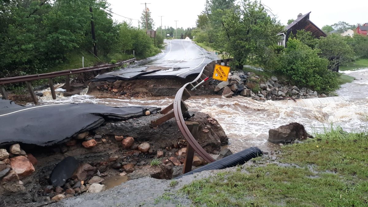 A whitewater stream flows through a broken road with twisted metal guardrails, broken and buckled pavement, and underlying eroded soil visible.