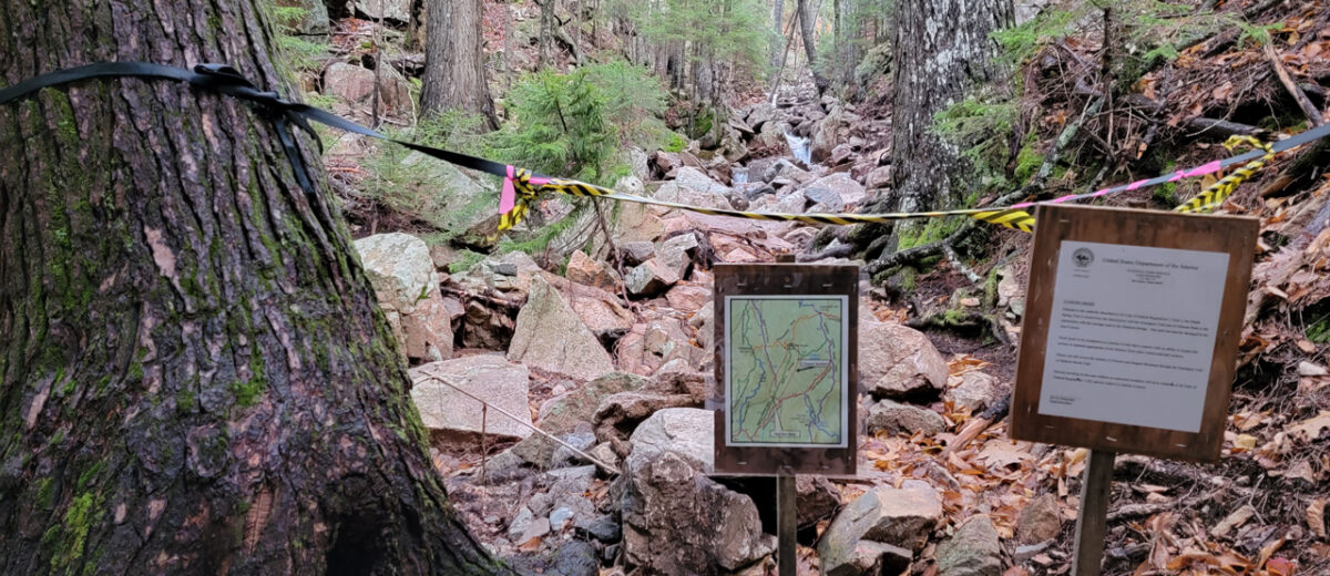 A view upstream along the Maple Spring Trail. A black rope with yellow caution tape is strung from a tree across the trail with signs indicating closure due to storm damage.