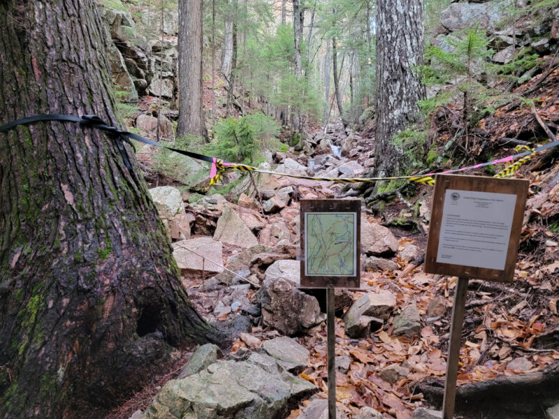 A view upstream along the Maple Spring Trail. A black rope with yellow caution tape is strung from a tree across the trail with signs indicating closure due to storm damage.