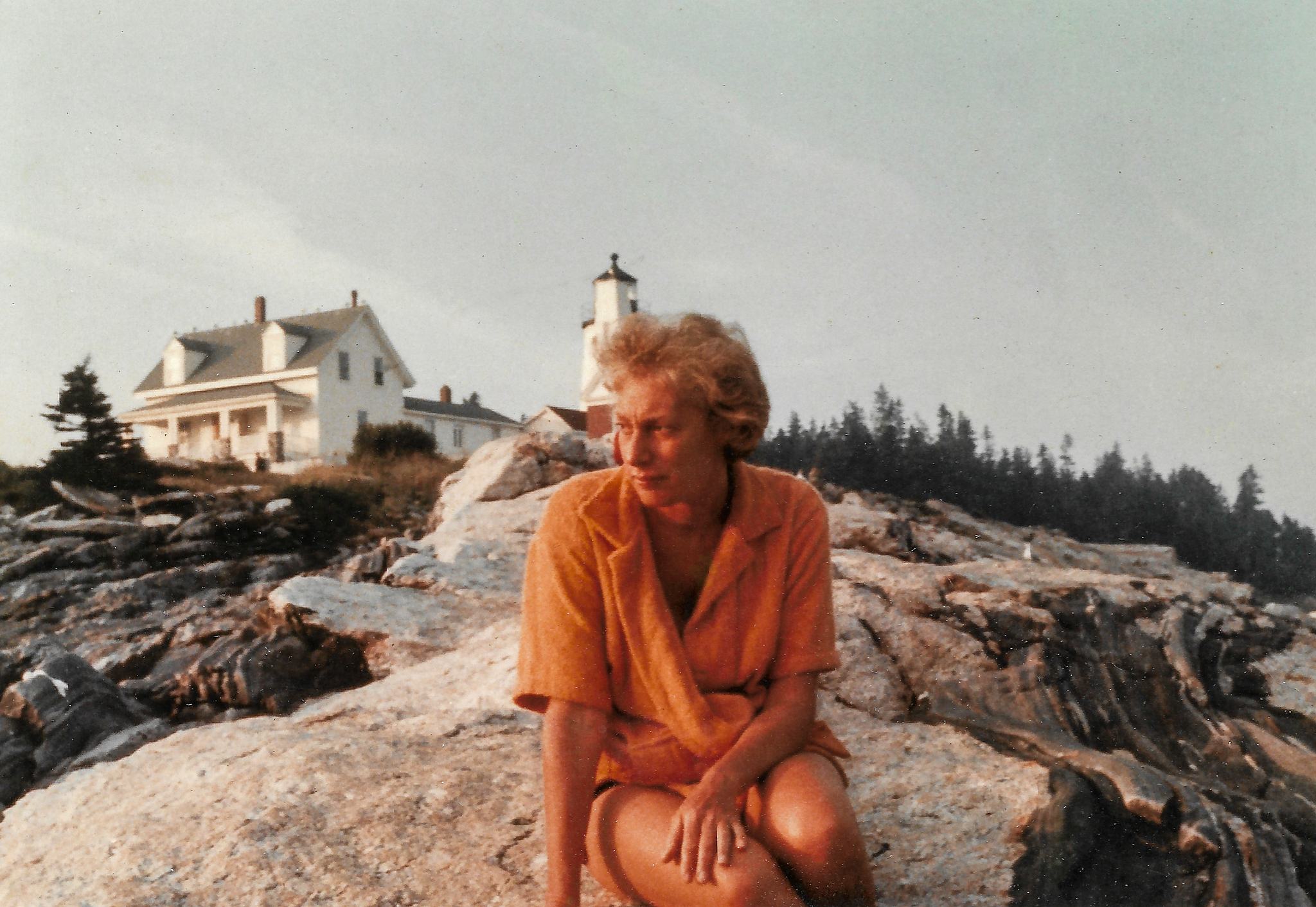 A person sits on rocks with white house and lighthouse in background.