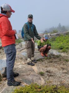Three people stand around an area of bare ground on the foggy summit of Cadillac Mountain.