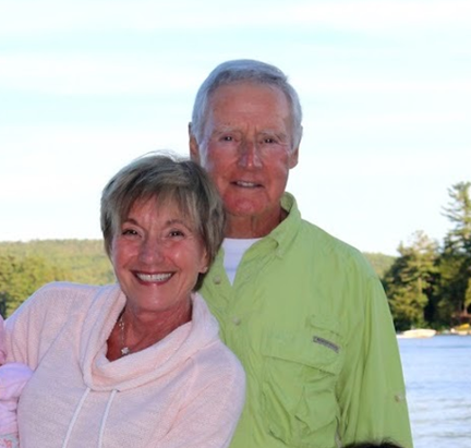 Cathy and Jim Gero face camera, smiling, with blue sky and water behind them in background