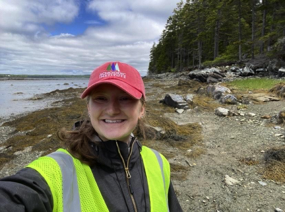 A smiling person wearing a Schoodic Institute ball cap and yellow safety vest takes a selfie photo on a rocky and seaweed covered shore with spruce forest in background