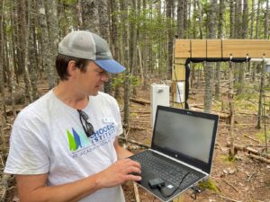 Peter holds a laptop as he stands in the forest next to the station, a metal cabinet mounted on a wooden post.