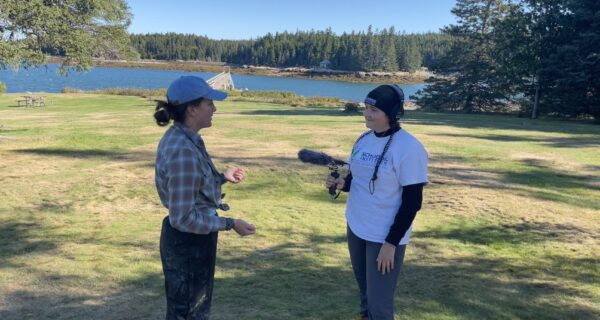 Olivia is interviewing Maya Peletier with her headphones and shotgun mic. The two are standing in a grassy area at Frazer Point with Mosquito Harbor in the background.