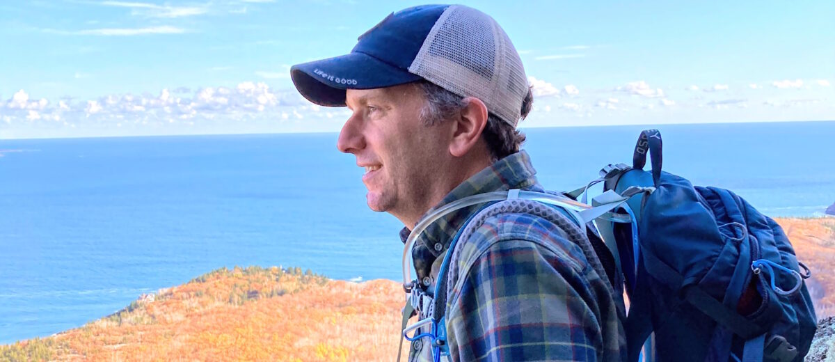A person wearing a baseball cap, plaid flannel shirt, and backpack leans on a rock and looks out across land and sea.