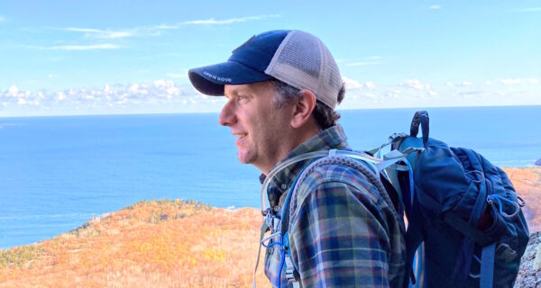 A person wearing a baseball cap, plaid flannel shirt, and backpack leans on a rock and looks out across land and sea.