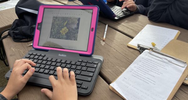A wooden picnic table is covered with clipboards, pencils, backpacks, and iPad tablets attached to keyboards. Students’ hands type at the keyboards.