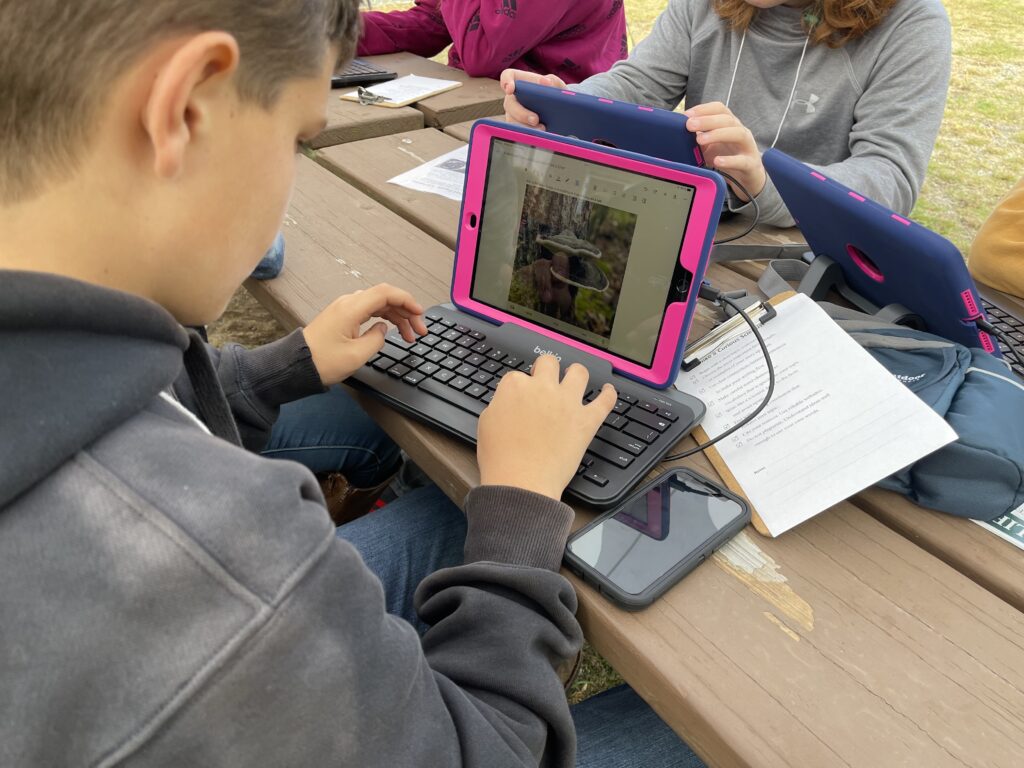 A middle school student sits at a picnic table and uses an iPad and attachable keyboard to type a paragraph. A photo of a lichen growing on a tree is in the document on the screen. 