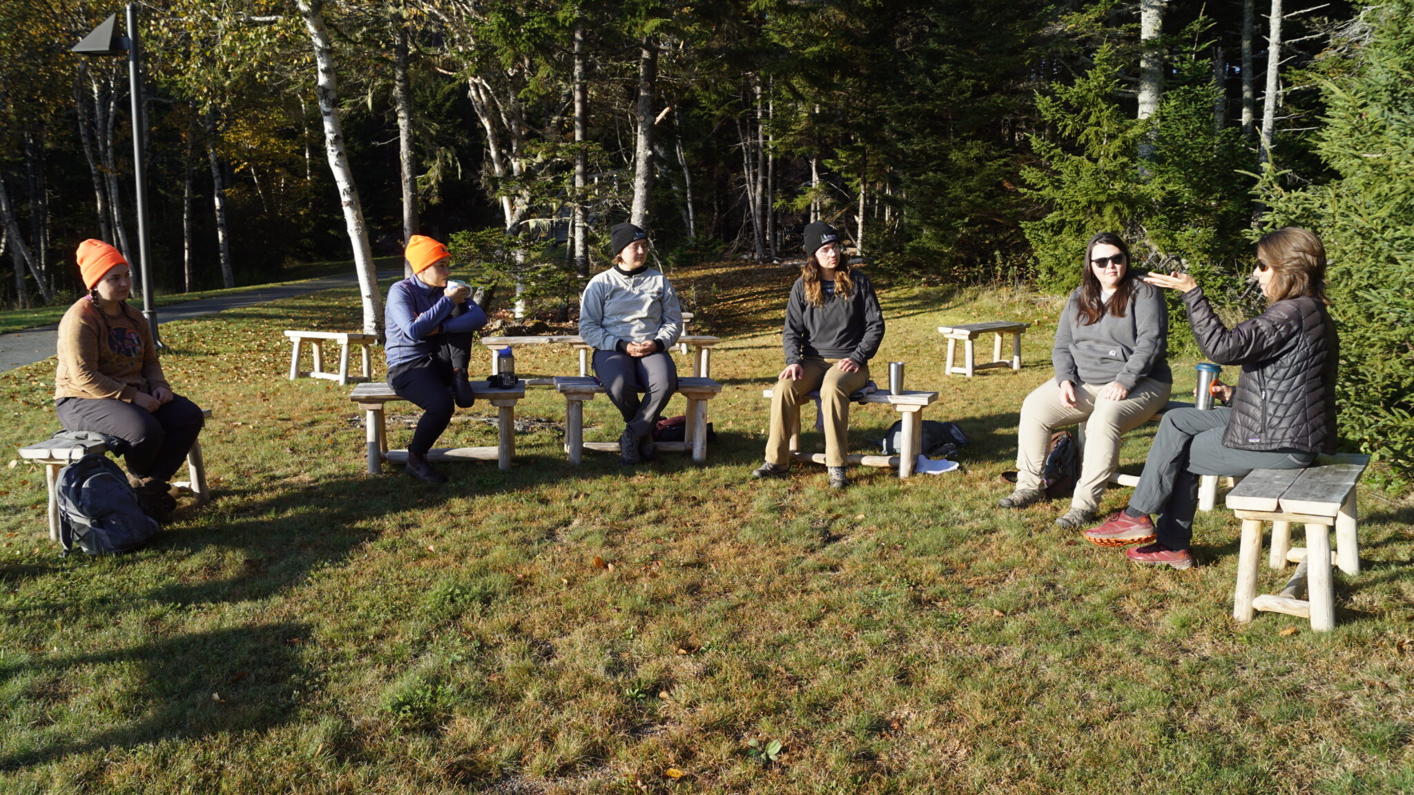 Group of people sitting outdoors on wooden benches in grassy area of Schoodic Institute campus.