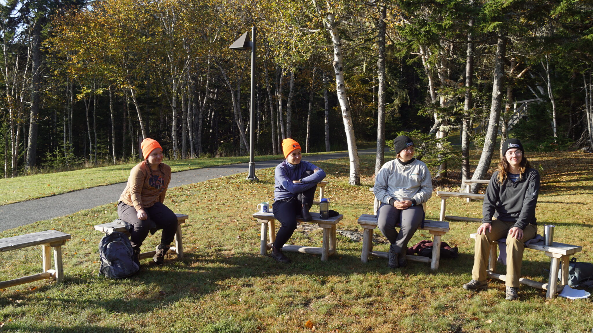 Group of four people sitting in outdoor classroom, which consists of homemade wooden benches in a circular position outdoors on grassy area.