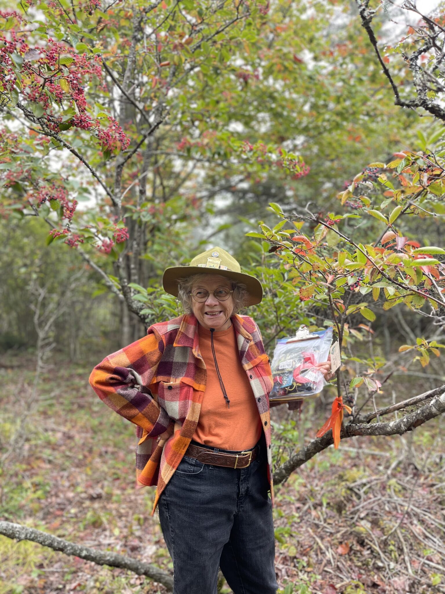 Priest stands in front of a study tree with her right hand on her hip, looping off to the right. In her left hand she carries a clipboard with a sealed plastic bag full of supplies.