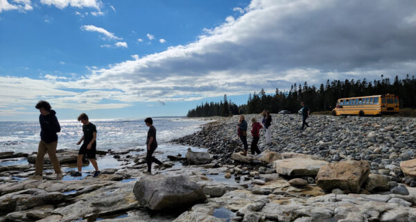 Group of high school students walk across rocky beach on Mount Desert Island, with view of horizon and ocean in background. To the left, a school bus is parked beyond a rocky beach.