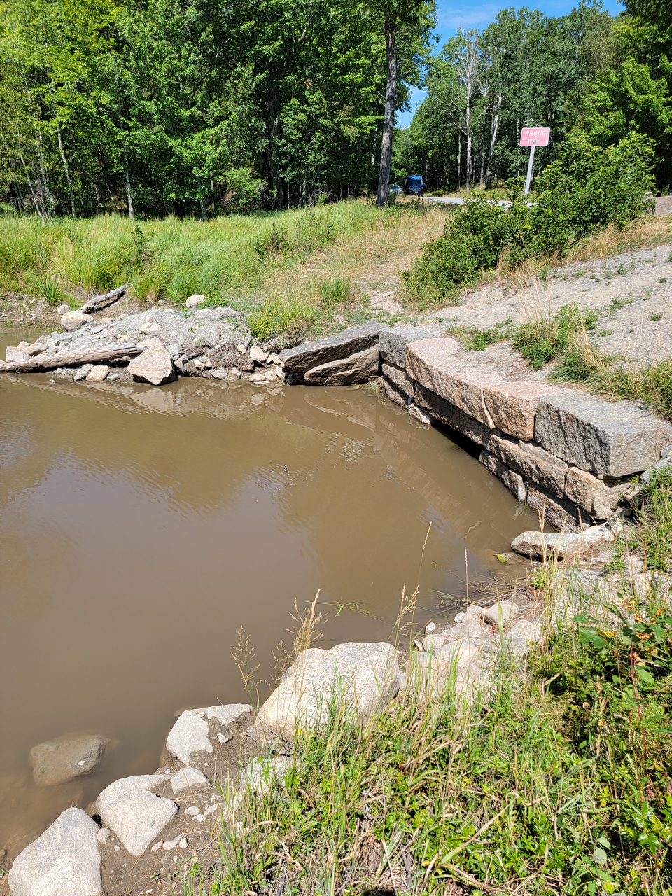 A pool of muddy water is blocked by a clogged stone culvert beneath a road.