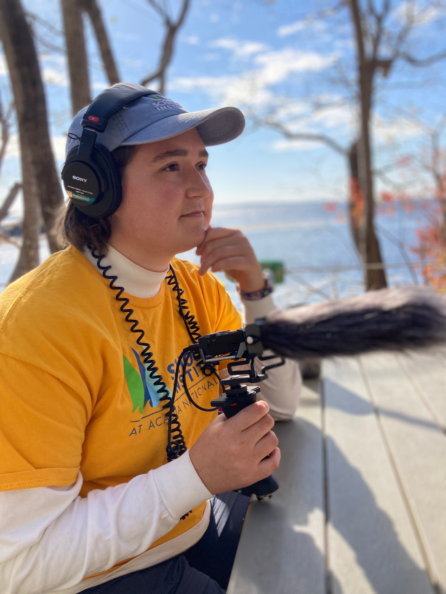 Olivia Milloway sits at an outside picnic table with podcast equipment as she interviews a podcast guest. The ocean can be seen behind her, under a bright blue sky.