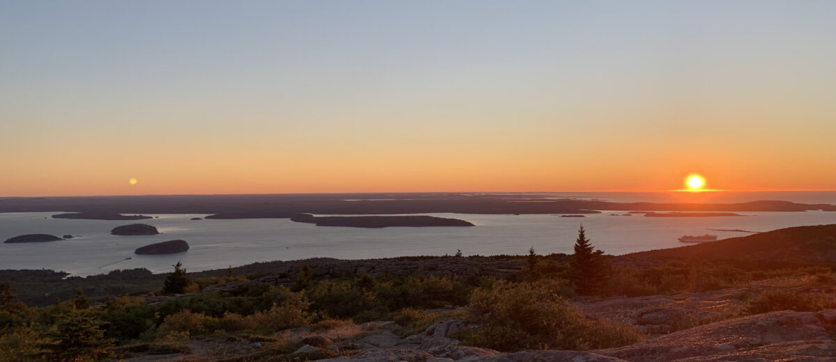 View of the sunrise from Cadillac Mountain in Acadia National Park. The sun is cresting the horizon line and islands are seen in the bay.