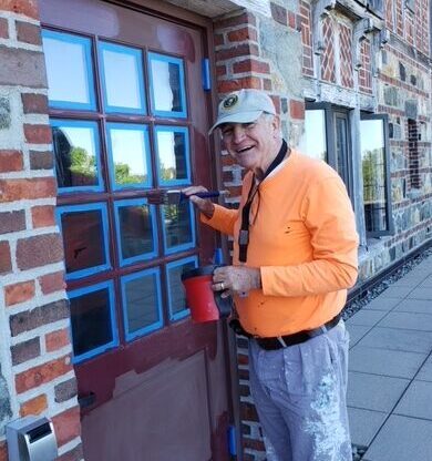 While smiling at the camera, volunteer host Tommy Jarrett applies a fresh coat of paint to an exterior door at Rockefeller Hall on the Schoodic Institute campus.