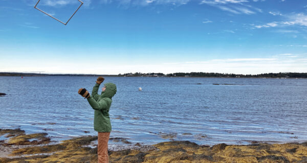 A Project ASCO participant throws a quadrant into the air haphazardly to help with measuring the amount of rockweed in a certain area. The ocean is shown in the background, under a blue sky.
