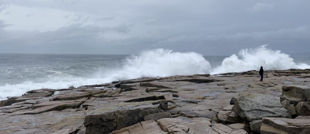 a lone figure stands on an expanse of rock and watches waves crash against the shore beneath a stormy sky