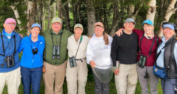 The 2021 Schoodic Notes volunteer group stand together with arms linked, smiling at the camera, with the forest shown behind them.