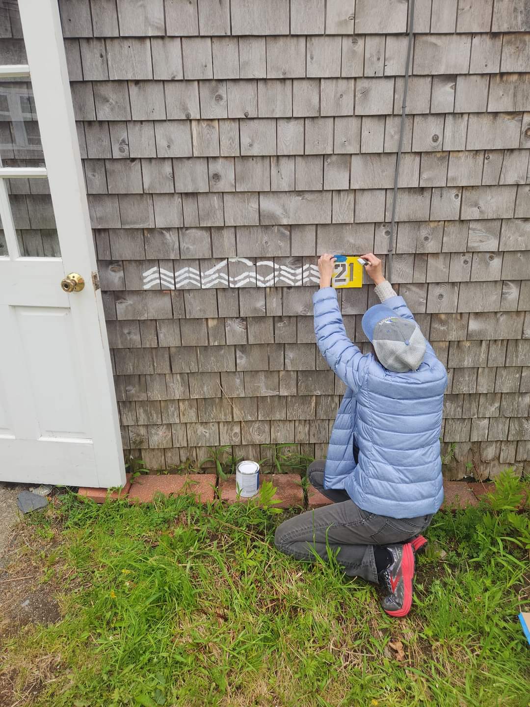 Catherine Schmitt uses a stencil to paint 2021 after a series of wavy lines four feet up the side cedar-shingled building.