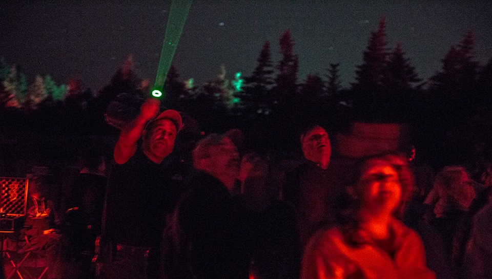 A crowd of people gaze up at the night sky, while one person holds a laser pointer toward the sky to direct attention as he teaches about the constellations.