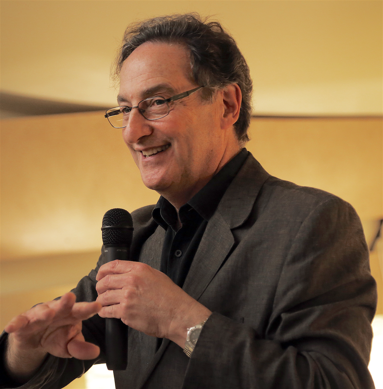 Headshot of Ira Flatow, renowned science communicator and public radio host. From a side-profile view, he is shown holding a microphone against a golden backdrop.