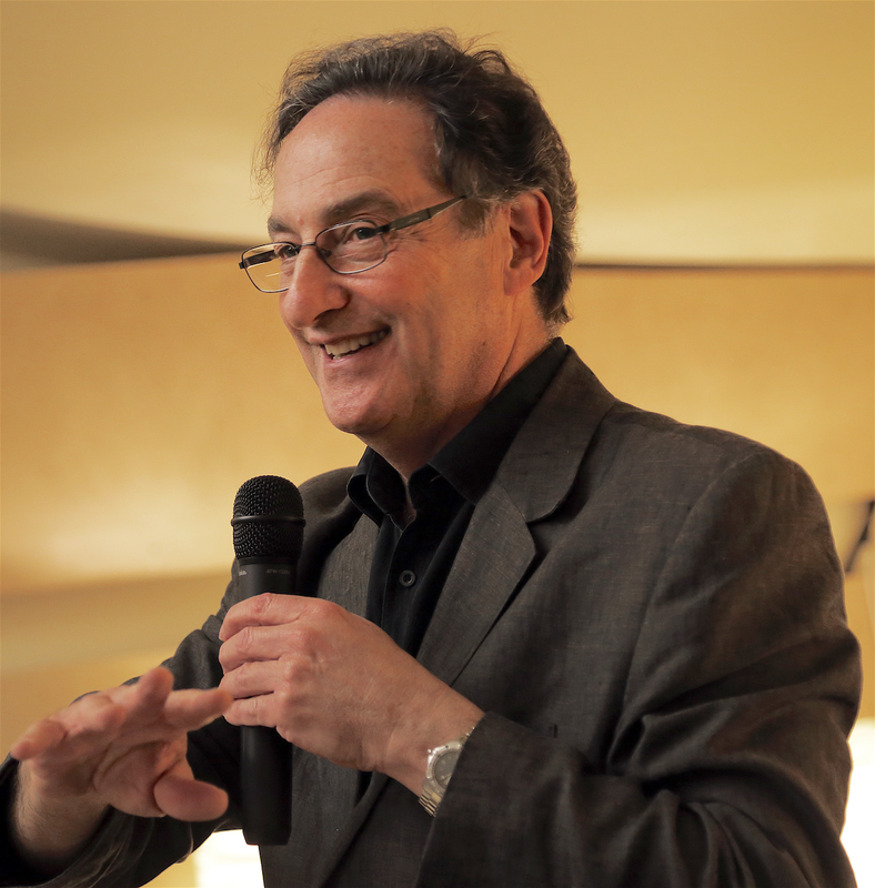 Headshot of Ira Flatow\, renowned science communicator and public radio host. From a side-profile view\, he is shown holding a microphone against a golden backdrop.