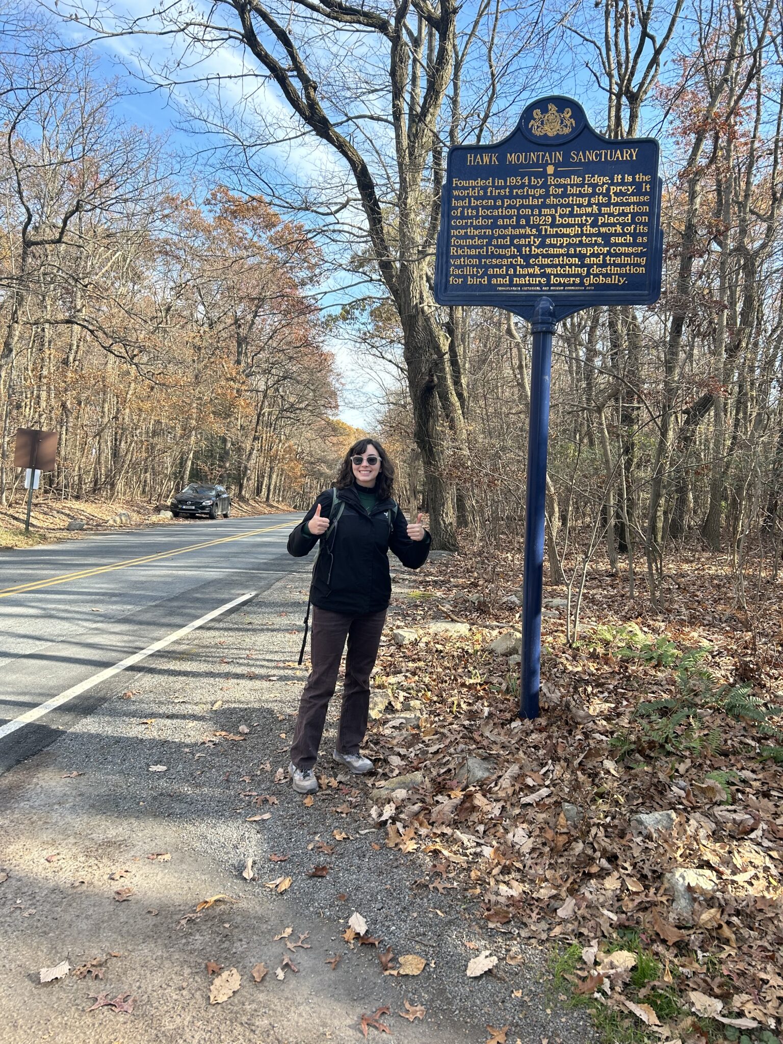 Brooke Goodman gives two thumbs up as she stands next to a roadside sign for the Hawk Mountain Sanctuary.