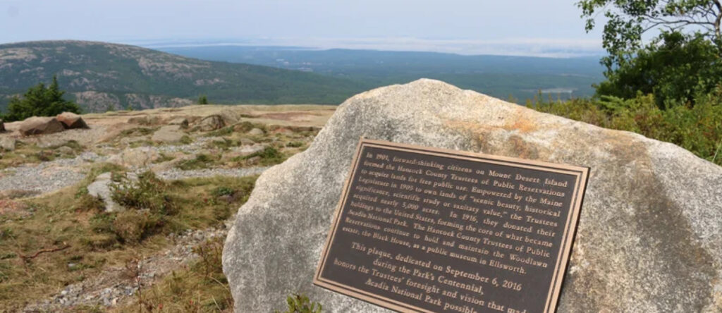 A plaque on a boulder in Acadia National park commemorates the foresight and vision that made the park possible. In the background are the rolling hills of Acadia, and, beyond, the open waters of the Atlantic.