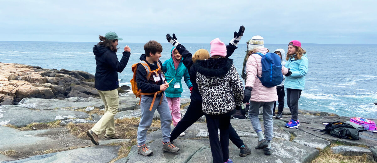 Standing on the rocky coast, a group of middle school students celebrate happily with arms in the air alongside an education fellow. The ocean is seen in the background.