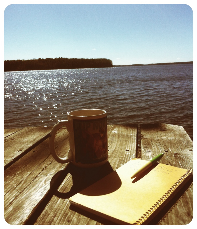 A close-up view of a coffee mug, journal, and pen sitting on a dock near the ocean. A sepia-toned filter is used, tinting the photo and giving it an antique-aesthetic. 
