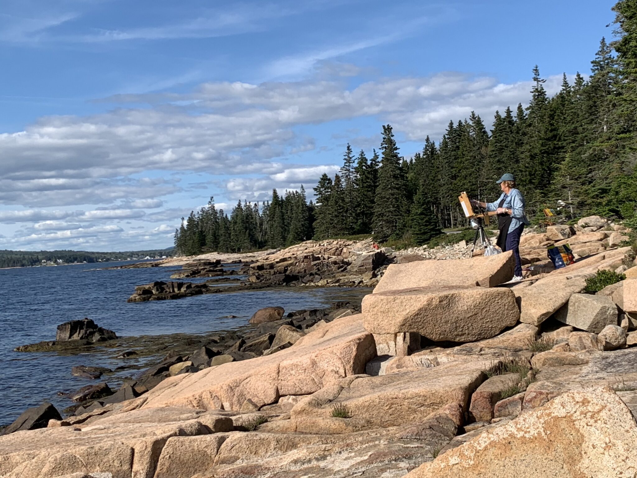 A painter stands in front of an easel stationed on the rocks along the Schoodic Peninsula coastline, while facing the open ocean and painting the scene.
