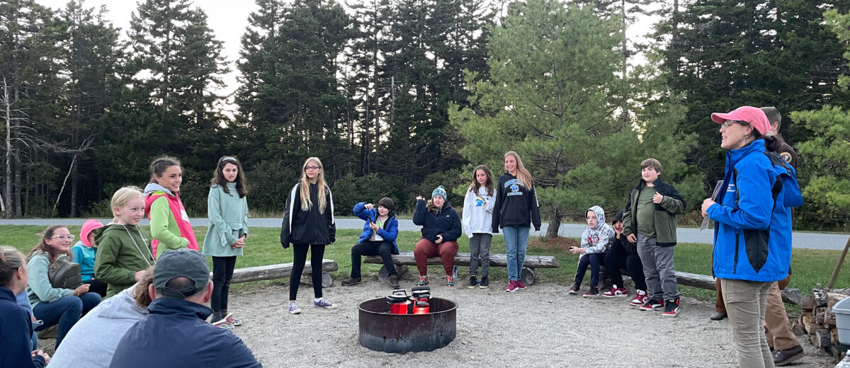 A group of middle school, students, teachers, and rangers gather around a campfire on the Schoodic Institute campus.
