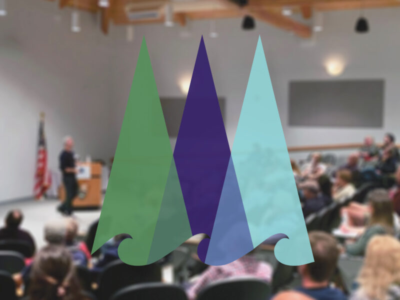 A crowded auditorium at Schoodic Institute listens to a presenter, standing at a podium. Overlaid is the Schoodic Institute logo artwork, depicting a stylized re-imagining of trees and ocean waves.