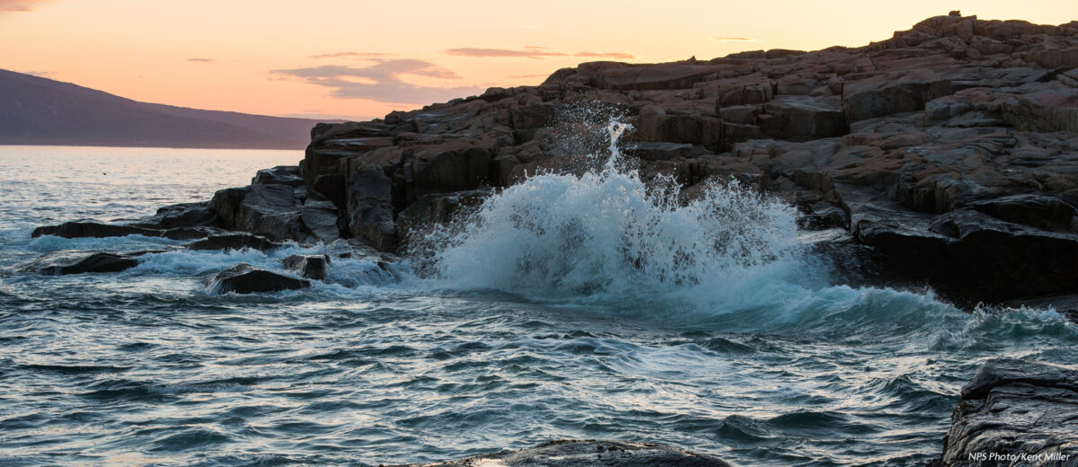 A wave crashes against the rocky coast on the Schoodic Peninsula at sunset.
