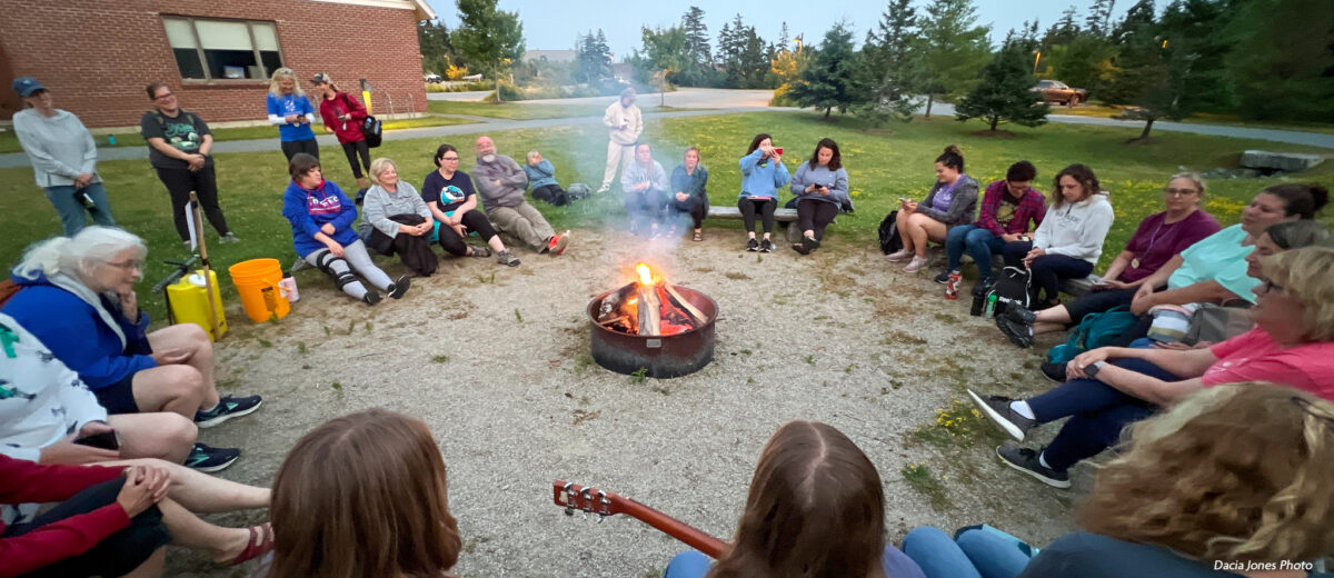 A group of students and visitors gather around a campfire on the Schoodic Institute campus.