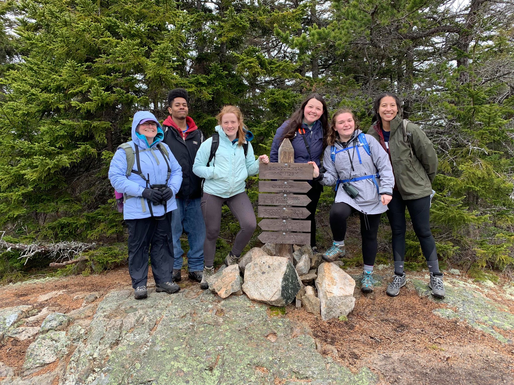 A group of hikers stand near a wooden summit post, smiling for the camera.